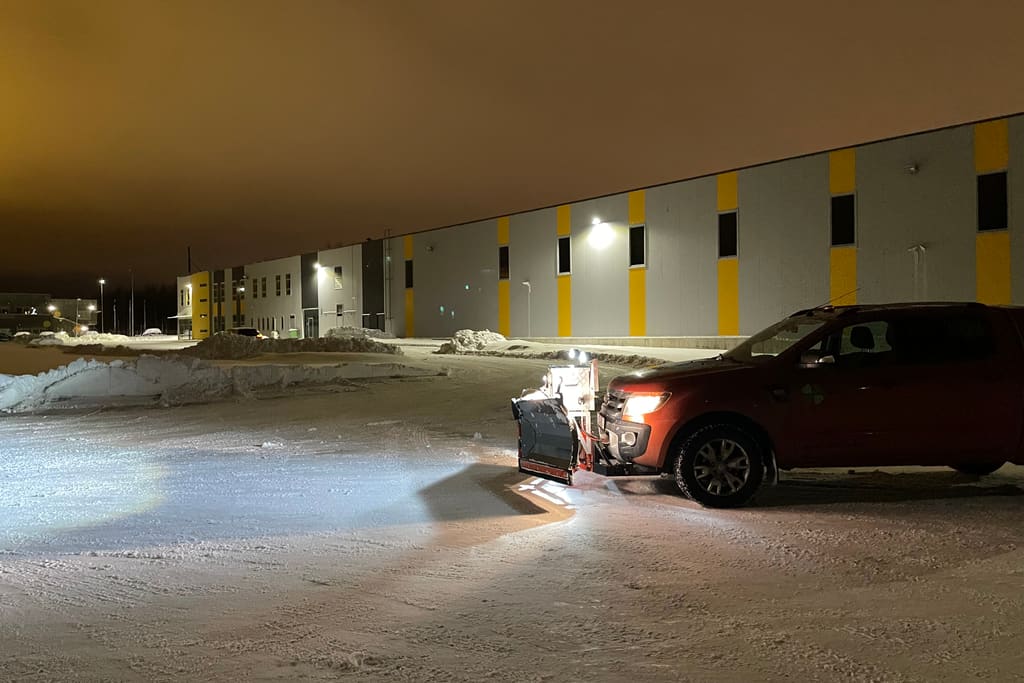 Sahaga pikap puhastab öösel laohoone parklat, esituled valgustavad puhastatud asfalti ja servas on lumevallid