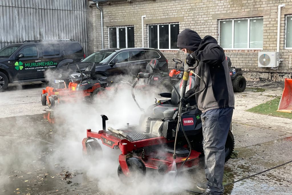 Töötaja peseb survepesuriga niidukit, ümberringi aurupilv, taustal Murumeeste sõidukid