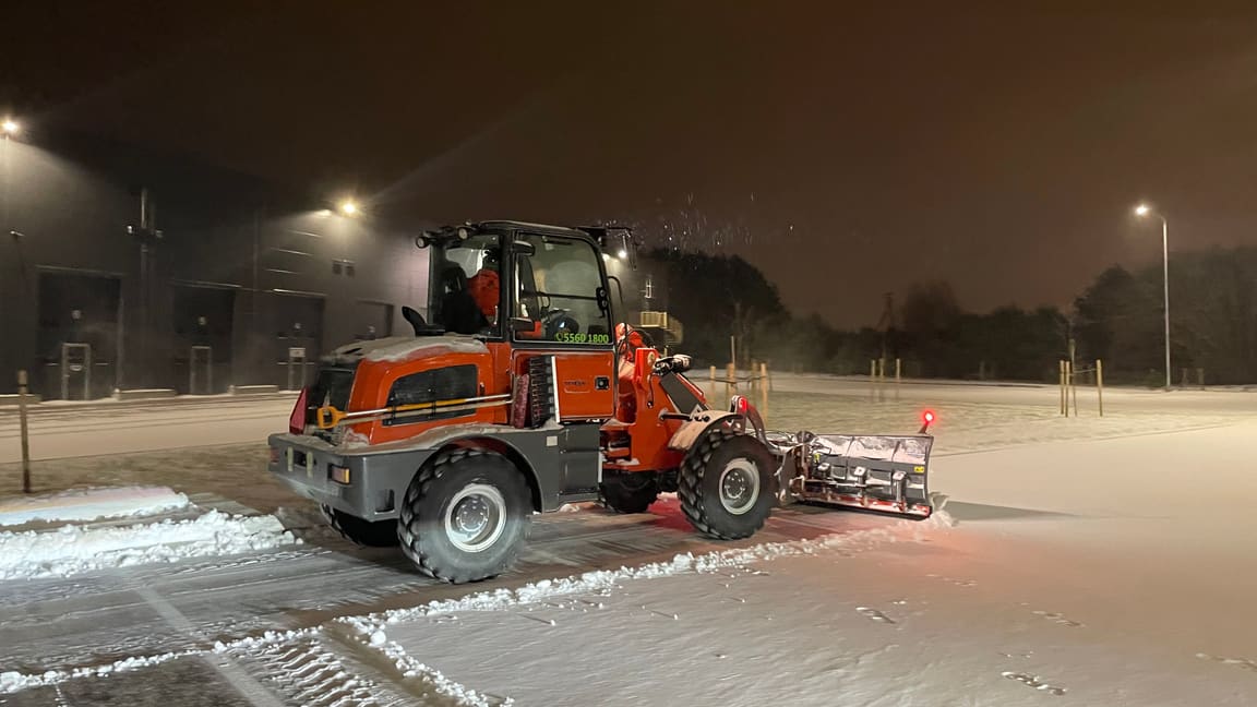 Rataslaadur lükkab öösel lumesajus ärihoone parklast lund, juht kabiinis